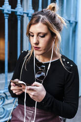 Young fashionable woman listening music while using mobile phone against window - JMPF00622