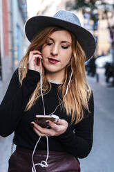 Blond woman wearing hat listening music through mobile phone while standing on street - JMPF00625