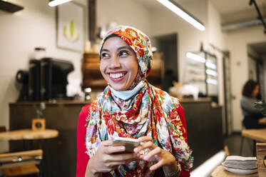 Smiling Muslim woman in floral hijab looking away while holding mobile phone at cafe - JCMF01601