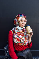 Lächelnde Frau hält Einweg-Kaffeebecher gegen schwarze Wand während COVID-19 - JCMF01606