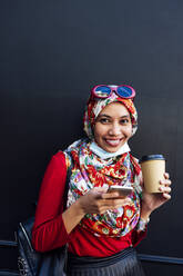 Lächelnde Frau mit Hidschab, die ein Handy und eine Kaffeetasse gegen eine schwarze Wand hält, während einer Pandemie - JCMF01607