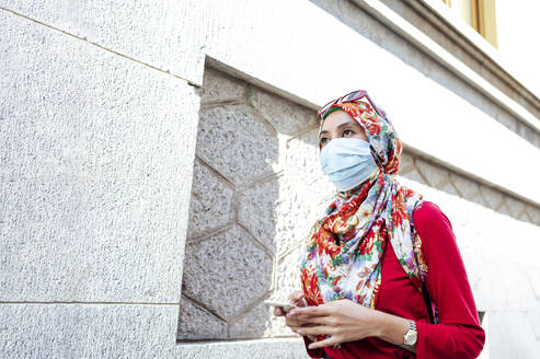 Frau mit Hidschab und Gesichtsschutz, die ihr Smartphone hält, während sie während der COVID-19 an einer Mauer vorbeiläuft - JCMF01612