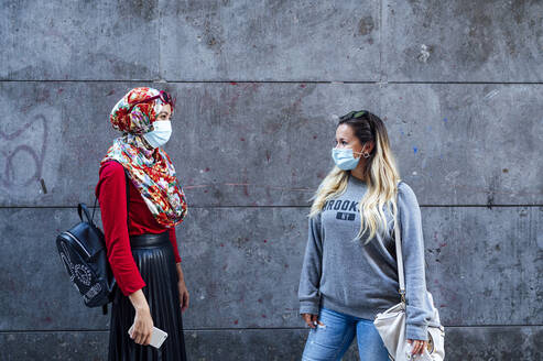 Female friends looking at each other while standing against gray wall during COVID-19 - JCMF01624