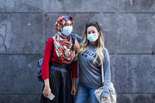 Woman wearing face mask while standing with hand on Muslim friend's shoulder against gray wall during pandemic - JCMF01625