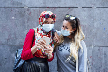 Muslimische Frau teilt Smartphone mit Freundin vor grauer Wand - JCMF01626