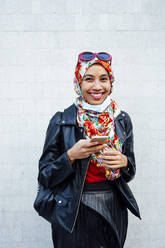 Smiling woman wearing leather jacket while holding mobile phone against wall during COVID-19 - JCMF01637