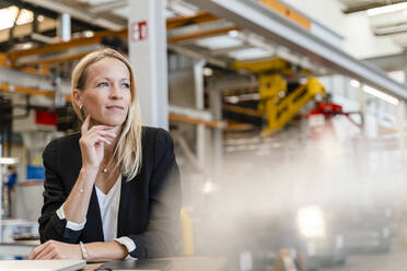Lächelnde nachdenkliche Geschäftsfrau mit Hand am Kinn in einer Fabrik sitzend - DIGF13265