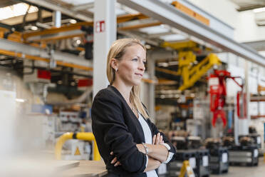 Blond businesswoman with arms crossed looking away while standing in factory - DIGF13266