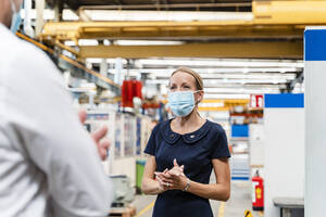 Female entrepreneur discussing with male colleague while standing in factory during pandemic - DIGF13271