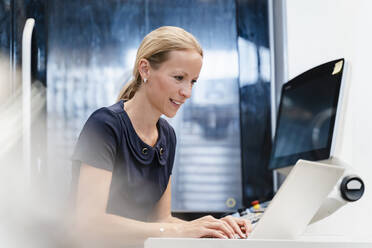 Lächelnde Geschäftsfrau, die an einem Laptop arbeitet, während sie in der Industrie steht - DIGF13279