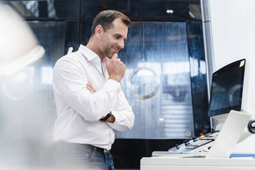 Lächelnder männlicher Unternehmer mit Hand am Kinn, der auf einen Laptop schaut, während er in einer Fabrik steht - DIGF13284