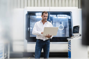 Businessman concentrating while working on laptop standing in factory - DIGF13285