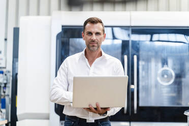 Confident businessman holding laptop while standing in industry - DIGF13286