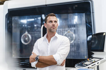 Confident businessman looking away with arms crossed while standing against door in factory - DIGF13292