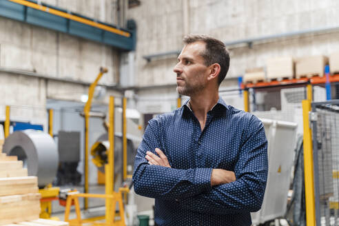 Nachdenklicher männlicher Unternehmer mit verschränkten Armen, der wegschaut, während er in einer Fabrik steht - DIGF13302