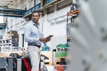 Male entrepreneur holding digital tablet looking away while standing in industry - DIGF13329