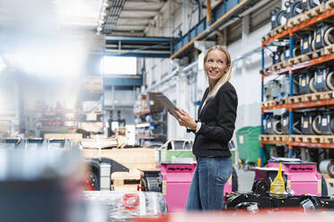 Glückliche Unternehmerin, die ein digitales Tablet hält und wegschaut, während sie in einer Fabrik steht - DIGF13335