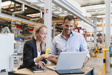 Smiling businessman and female entrepreneur using laptop at factory - DIGF13347