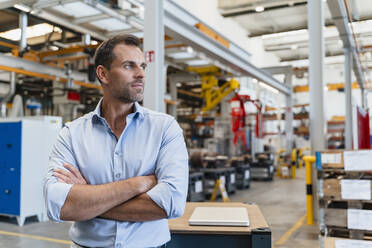 Confident male entrepreneur with arms crossed looking away while standing in factory - DIGF13357