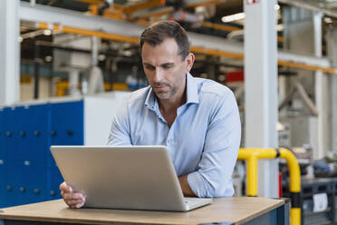 Männlicher Unternehmer, der einen Laptop benutzt und sich auf einen Schreibtisch in einer Fabrik stützt - DIGF13359