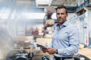 Confident businessman holding digital tablet while standing at factory - DIGF13364