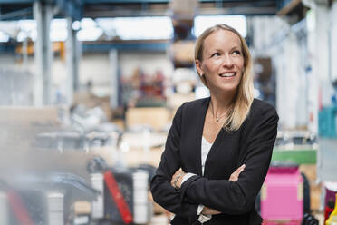 Smiling thoughtful businesswoman with arms crossed standing in factory - DIGF13366