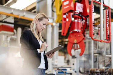 Blonde Geschäftsfrau, die ein digitales Tablet benutzt, während sie an einer Klauenmaschine in einer Fabrik steht - DIGF13385