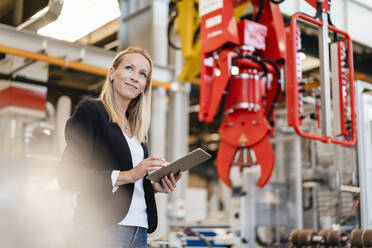 Lächelnde blonde Geschäftsfrau, die wegschaut und ein digitales Tablet in der Hand hält, in einer Fabrik stehend - DIGF13386