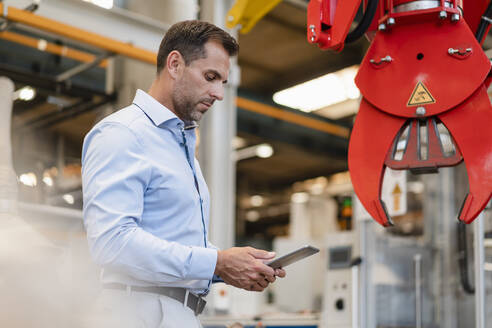 Männlicher Unternehmer, der ein digitales Tablet benutzt, während er vor einer Klauenmaschine in der Industrie steht - DIGF13394