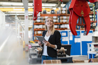 Lächelnde Geschäftsfrau, die eine Klaue untersucht und ein digitales Tablet in einer Fabrik hält - DIGF13401