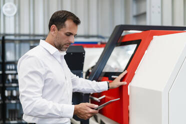 Männlicher Unternehmer, der ein digitales Tablet benutzt, während er in einer Fabrik vor einer Maschine steht - DIGF13403