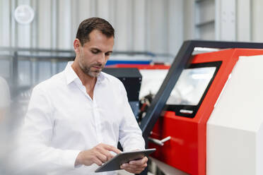 Geschäftsmann, der ein digitales Tablet benutzt, während er an einer Maschine in einer Fabrik steht - DIGF13404