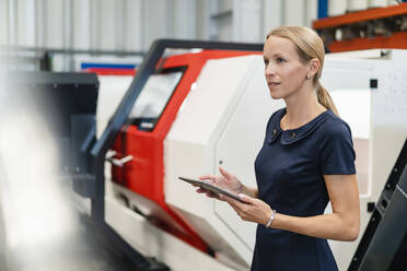 Blonde Geschäftsfrau mit digitalem Tablet vor Maschinen in der Industrie - DIGF13407