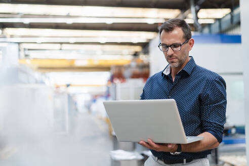 Geschäftsmann, der konzentriert an einem Laptop in einer Fabrik arbeitet - DIGF13415