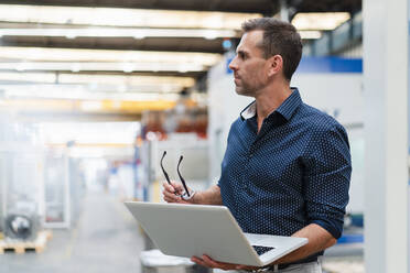 Male entrepreneur holding laptop looking away while standing in factory - DIGF13417