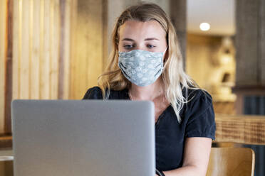 Junge blonde Frau mit blauer Gesichtsmaske, die am Tisch sitzt und einen Laptop benutzt. - MINF15251