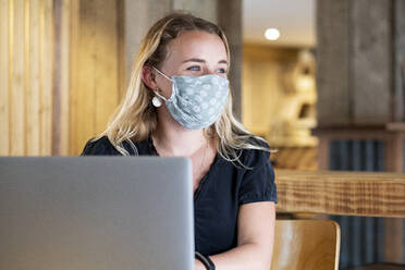 Junge blonde Frau mit blauer Gesichtsmaske, die am Tisch sitzt und einen Laptop benutzt. - MINF15252