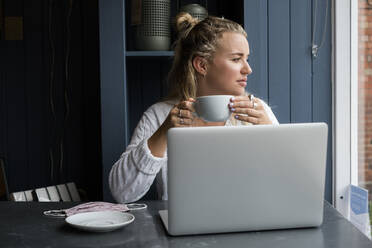 Junge blonde Frau, die allein an einem Cafétisch mit einem Laptop-Computer sitzt und aus der Ferne arbeitet. - MINF15277