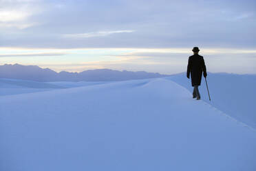Rückansicht eines Mannes mit Mantel und Melone, der mit einem Stock durch eine verschneite Landschaft geht. - MINF15282