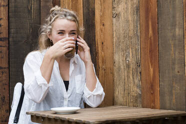 Junge blonde Frau, die allein in einem Café sitzt, ein Mobiltelefon benutzt und aus der Ferne arbeitet. - MINF15300