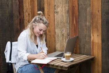 Junge blonde Frau sitzt allein an einem Cafétisch mit einem Laptop-Computer - MINF15302