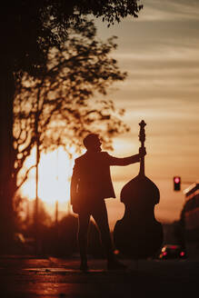 Silhouette eines Musikers, der einen Kontrabass hält und bei Sonnenuntergang auf der Straße steht - GMLF00845