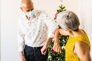 Positive mature couple in smart casual outfits and face masks greeting by bumping elbows while standing near decorated shiny Christmas tree - ADSF17639