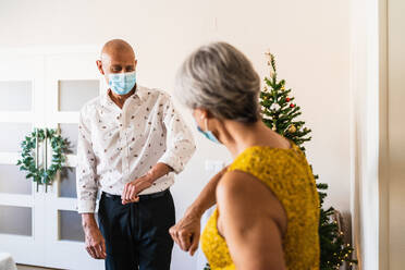 Positive mature couple in smart casual outfits and face masks greeting by bumping elbows while standing near decorated shiny Christmas tree - ADSF17644