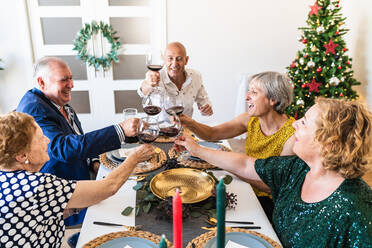 Glückliche reife Freunde versammeln sich an einem festlichen Tisch in der Nähe des leuchtenden Weihnachtsbaums und stoßen mit Weingläsern an, während sie gemeinsam den Weihnachtsabend feiern - ADSF17653