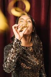 Female in glittering dress standing on red background and blowing party horn while having fun during holiday - ADSF17675