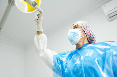 Zahnarzt in Arbeitsschutzkleidung beim Einstellen einer elektrischen Lampe in der Klinik während Covid-19 - JCMF01651