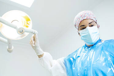 Dental doctor adjusting illuminated electric lamp while standing at clinic during Covid-19 - JCMF01652