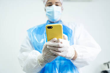 Female orthodontist wearing protective workwear using mobile phone while standing in clinic during Covid-19 - JCMF01654