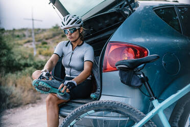 Selbstbewusste Radfahrerin mit Helm und Sonnenbrille, die im Kofferraum eines Autos sitzt und sich auf die Fahrt vorbereitet, während sie die Stiefel anzieht und in die Kamera schaut - ADSF17704